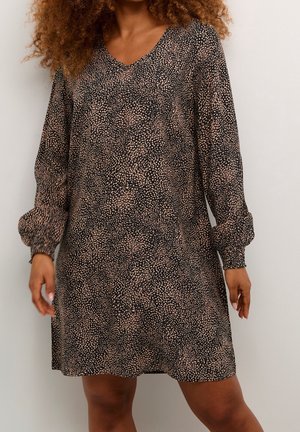 Kvinde iført en langærmet, V-hals kjole med sort og beige lille plettet mønster, stående mod en ensfarvet baggrund.