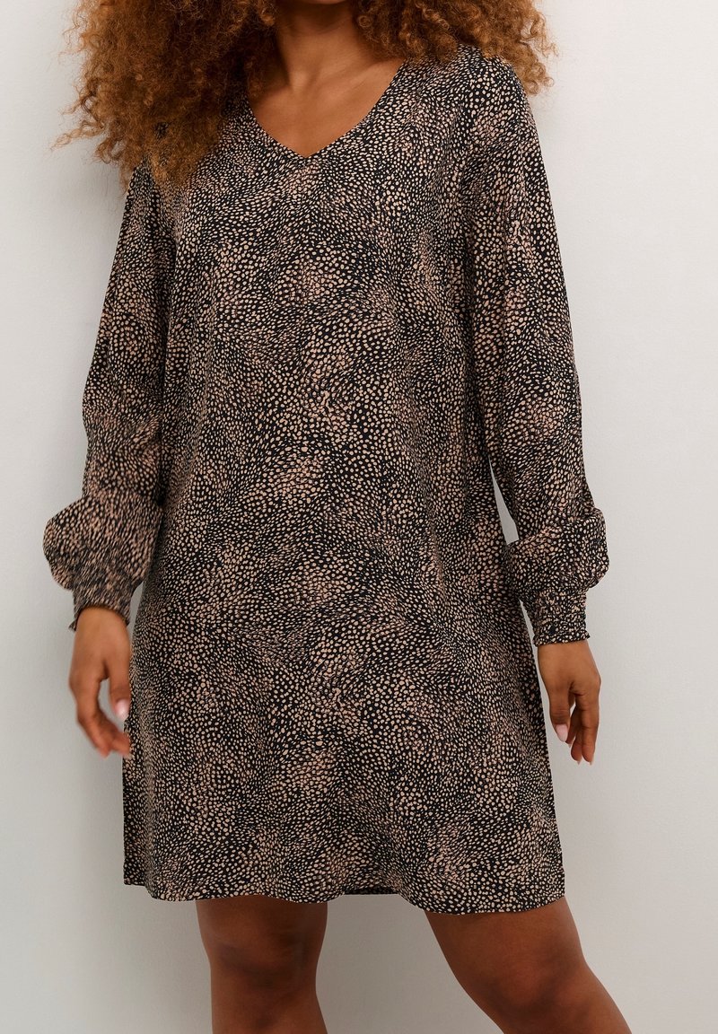 Femme portant une robe à manches longues avec un col en V et un petit motif tacheté noir et beige, debout devant un fond uni.