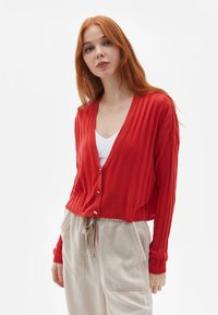Rød ribbet cardigan med V-udskæring, der har tre knapper og lange ærmer, parret med beige bukser, glat stofstruktur.