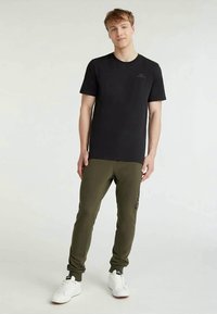 Jovem em pé, vestindo uma t-shirt preta, calças de treino verde-oliva com logotipo lateral e ténis brancos, contra um fundo claro simples.