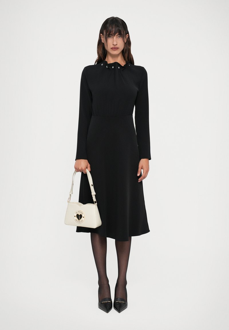 Donna in un abito midi lungo a maniche nere tiene in mano una piccola borsa bianca, indossando collant neri trasparenti e tacchi a punta neri.