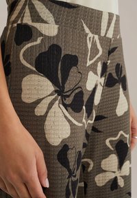 Hlače z cvetličnim vzorcem v teksturiranem, tkanem materialu. Barve vključujejo zemeljske tone z velikimi črnimi in kremnimi cvetovi. Opredeljen pas in žepi.