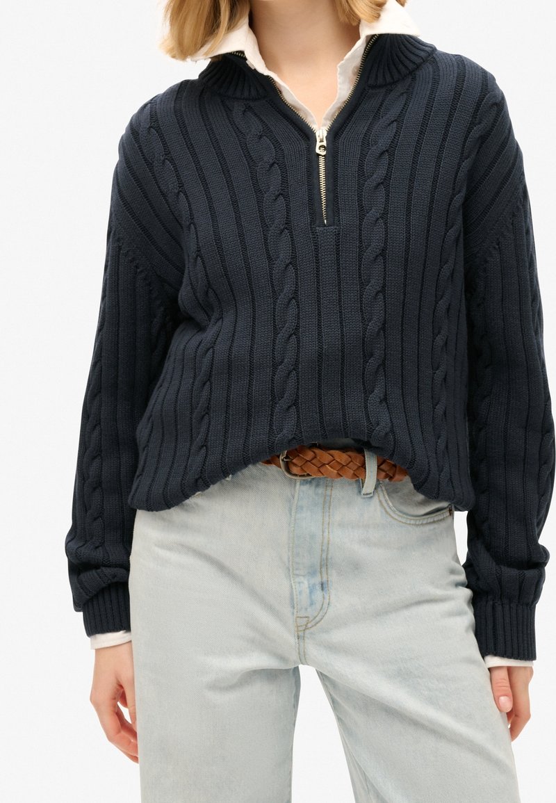 Marineblauer Strickpullover mit halbem Reißverschluss und Zopfmuster. Kombiniert mit hellblauen Jeans und einem geflochtenen braunen Gürtel.