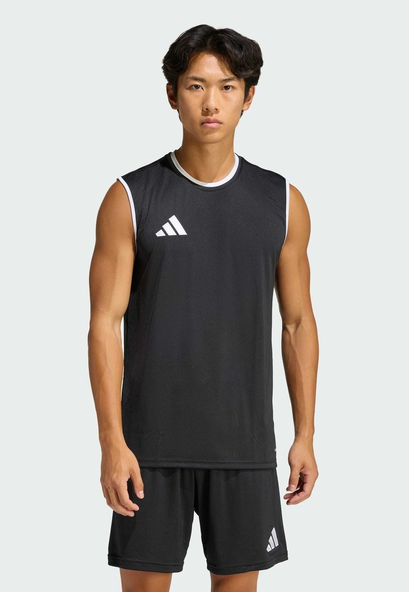 Jonge man die een zwart mouwloos sportshirt en bijpassende korte broek draagt met witte Adidas-logo’s, staand tegen een effen lichte achtergrond.