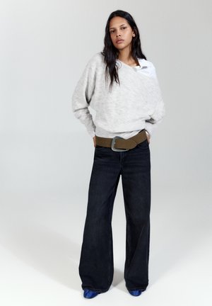 Femme aux longs cheveux foncés portant un pull gris clair, un jean large foncé, une ceinture marron et des chaussures bleues, debout les mains dans les poches.