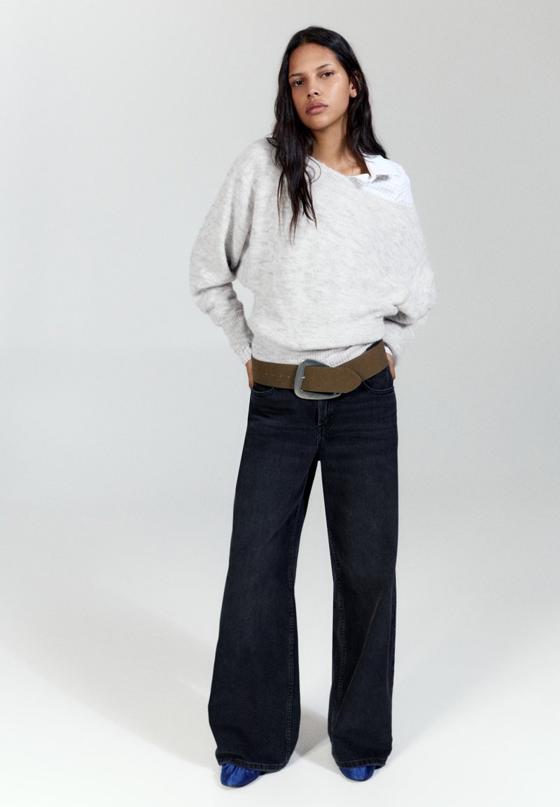 Femme aux longs cheveux foncés portant un pull gris clair, un jean large foncé, une ceinture marron et des chaussures bleues, debout les mains dans les poches.