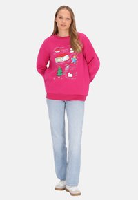 Fukszia pulóver játékos ünnepi grafikákkal, köztük karácsonyi témájú illusztrációkkal. Világoskék, bővülő szabású farmernadrággal és fehér sportcipővel párosítva.