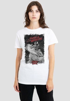 Young woman wearing white t-shirt with black-and-white dancing woman graphic and red French text "La famille est un monstre."