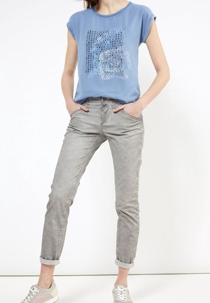 Světle modrý top s krátkým rukávem a grafickým potiskem; doplněno o šedé zvednuté džíny; modelka má na nohou béžové tenisky. Jednoduchý design, měkká textura.