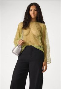 Woman with long dark hair wears sheer yellow textured top, black pants with pockets, and holds small silver handbag over shoulder.