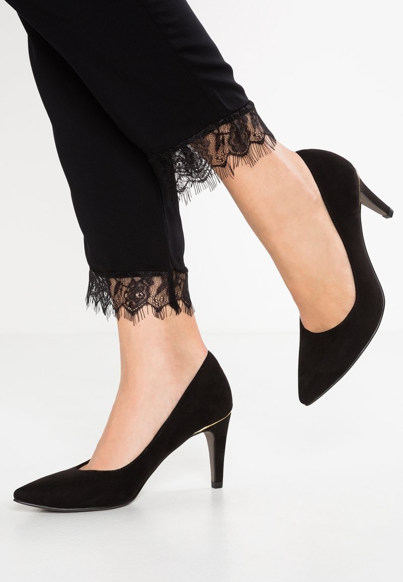 Jambes portant un pantalon noir orné de dentelle et des chaussures à talons pointues en daim noir, avec un talon levé et un pied à plat.