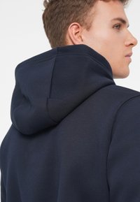Felpa blu navy caratterizzata da un tessuto morbido e texturizzato, con cappuccio strutturato e una vestibilità comoda, arricchita da dettagli di cucitura visibili.