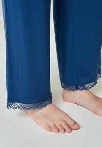 Siniset pyjamahousut, joissa on sileä pinta ja herkät kehysreunat helmassa, yhdistettynä paljaisiin jalkoihin vaalealla taustalla.