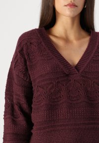 Vinröd stickad tröja med V-ringning, med texturerade mönster och lös passform, som visar på detaljerade inslag i tyget.