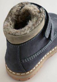 Tmavomodré semišové botas s šedým vnútorným podšívaním, suchým zipsom a bielym prešívaním pozdĺž podrážky. Štruktúrovaný povrch okolo goliera.