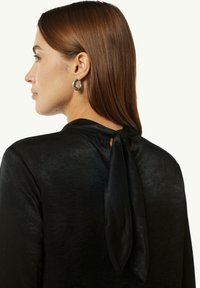 Schwarze Bluse mit weicher Textur, die auf der Rückseite einen Bindeschluss und eine Schlüsselöffnungsdetails aufweist. Große silberne Creolen sind sichtbar.