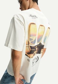 Witte katoenen T-shirt met een afbeelding op de achterkant die vliegtuigramen toont met een zonsondergangszicht, vergezeld van tekst en accenten.