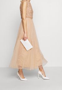 Helmikoristeltu vaaleanpunainen crop-top beige tylli-hameen kanssa, varustettuna valkoisella clutch-laukulla ja valkoisilla kärkitossuilla. Sileät, kerrokselliset tekstuuri.