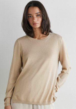 Kvinde med skulderlangt mørkt hår iført en beige langærmet sweater, stående mod en ensartet lys baggrund.