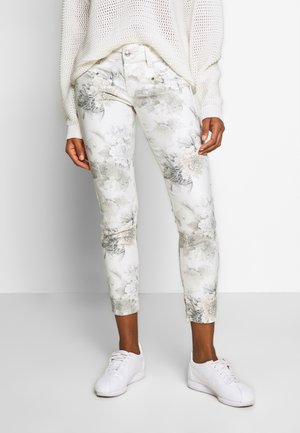 Jean à imprimé floral en blanc avec des tons gris clair et beige, présentant une coupe slim, un design à cinq poches et des accents en métal argenté. Associé à des baskets blanches.