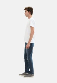 Una camiseta blanca de manga corta combinada con jeans azul oscuro y zapatillas grises, exhibida en vista lateral sobre un fondo liso.