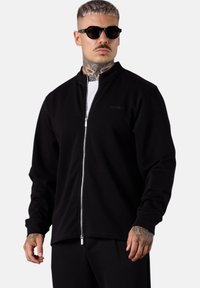 Schwarze Zip-Jacke aus weichem Stoff mit hohem Kragen, unsichtbaren Nähten, langen Ärmeln und slim fit Schnitt.