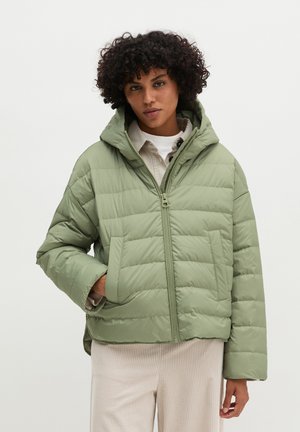 Vrouw met krullend haar die een lichtgroene donsjas met capuchon draagt over een wit hemd en lichte broek tegen een effen achtergrond.