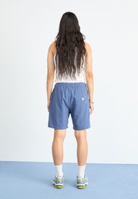 Blå shorts laget av pustende stoff, med elastisk midje, baklomme, og en liten logo på baksiden. Bært med hvite sokker og joggesko.