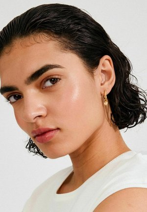 Gouden oorring met structuurdetails gedragen aan het linkeroor; kort, nat uitziend donker haar; gladde huid met subtiele highlight. Witte top.