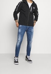 Black zip-up jacket with hood, featuring white side stripes and a logo. Worn with distressed blue jeans and black high-top sneakers.