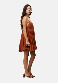 Rostfarbenes, ärmelloses Kleid mit Falten, fließendem Rock, weicher Textur und A-Linie-Form. Kombiniert mit braunen Blockabsatzsandalen.