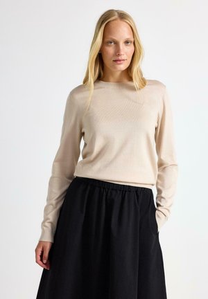 Blondynka ma na sobie beżowy sweter z długim rękawem oraz czarną spódnicę z elastyczną talią, stoi na jednolitym jasnym tle.