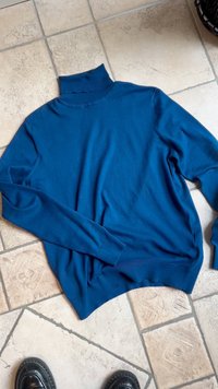 Maglione a collo alto lavorato a maglia blu, con maniche lunghe, dettagli a costine sul colletto e sull'orlo, texture morbida, posato su una superficie piastrellata.