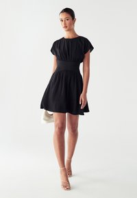 Black short dress with a fitted waist, short sleeves, and a flared skirt. Smooth fabric with no visible patterns, paired with strappy sandals.