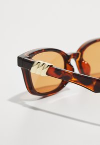 Ochelari de soare tortoiseshell maronii cu rame unghiulare, cu detalii aurii pe brațele ochelarilor și lentile maronii. Finisaj neted și lucios.