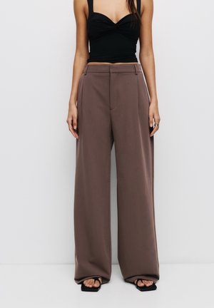Mujer con pantalones marrones de cintura alta y pierna ancha, top corto negro sin mangas y sandalias negras con puntera cuadrada y anillos en los dedos.