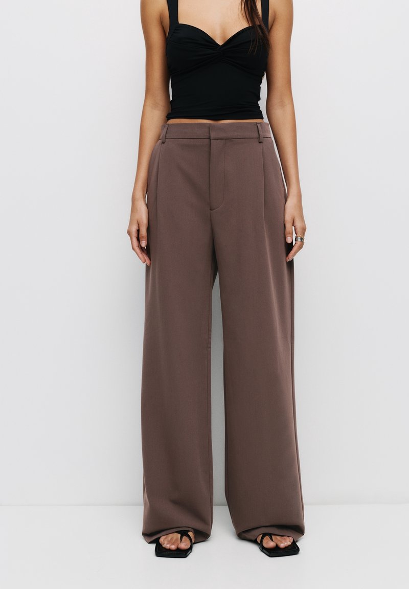 Femme portant un pantalon marron taille haute à jambes larges, un top court noir sans manches et des sandales noires à bout carré avec des bagues aux doigts.