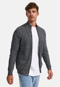 Chaqueta de punto gris con cuello alto, cierre de cremallera y mangas largas, llevada sobre una camiseta blanca y combinada con jeans de color azul claro.
