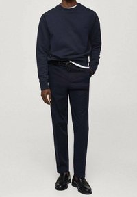 Man wearing black crewneck sweatshirt, black trousers with belt, black leather loafers, and white undershirt, standing with one hand in pocket.