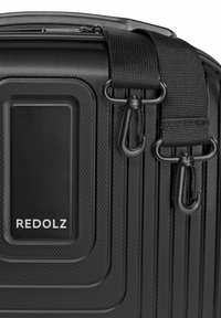 Zwarte gestructureerde harde koffer met afneembare schouderriem en metalen sluitingen, met een rechthoekig merklabel met "REDOLZ."