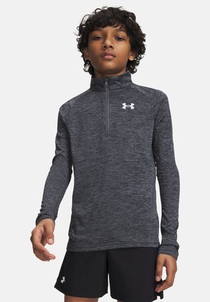 Young person with curly hair wearing a long-sleeve dark grey quarter-zip top and black shorts with the Under Armour logo.