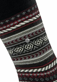 Calcetín de punto con un diseño de múltiples patrones en rojo, gris y negro, con una etiqueta metálica circular y motivos geométricos y florales en contraste.