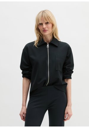 Frau mit blonden Haaren, die eine schwarze Jacke mit Reißverschluss und Tasche sowie passende schwarze Hose trägt, steht vor einem schlichten hellen Hintergrund.