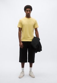 Κίτρινο βαμβακερό t-shirt, μαύρο σορτς μέχρι το γόνατο, λευκές κάλτσες, κρεμ αθλητικά παπούτσια. Το μοντέλο κρατάει ένα μαύρο σακάκι. Απλός σχεδιασμός, άνετη εφαρμογή.