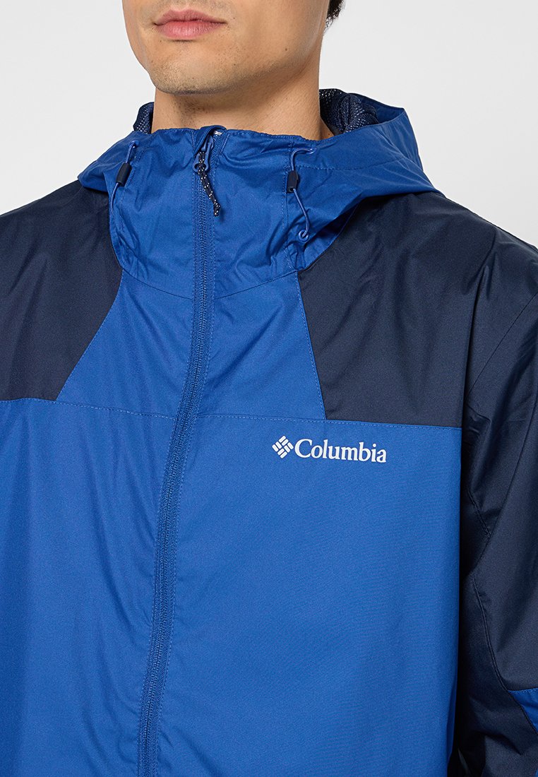 Veste imperméable bleue et marine avec une fermeture éclair sur le devant, capuche ajustable et logo sur la poitrine. Tissu léger avec une texture lisse.