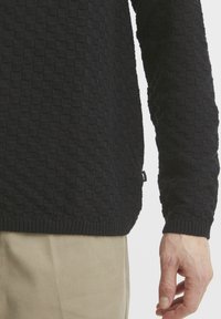 Black knitted sweater with a textured checker pattern and ribbed cuffs. Lightweight fabric. Side tag visible near hem.