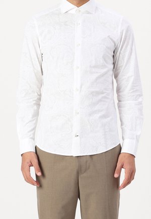 Homme portant une chemise blanche cintrée à manches longues avec des motifs floraux subtils et un pantalon beige, debout devant un fond blanc.