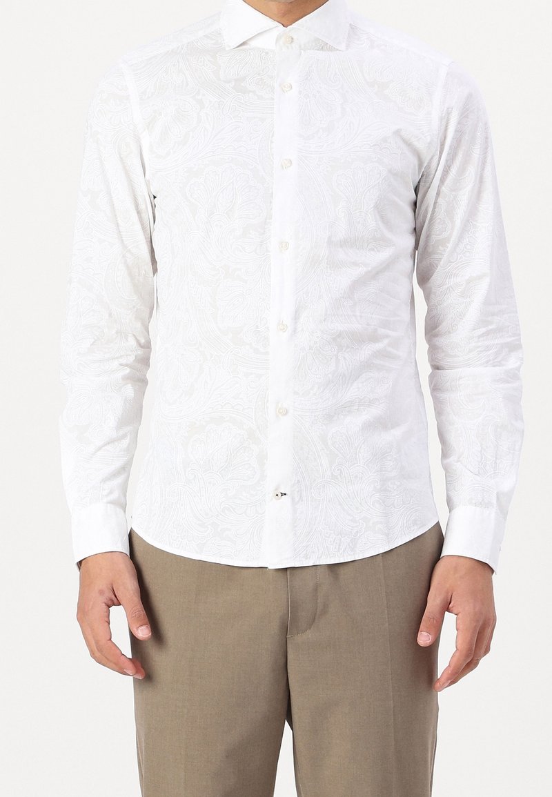 Homme portant une chemise blanche cintrée à manches longues avec des motifs floraux subtils et un pantalon beige, debout devant un fond blanc.