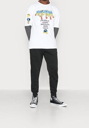 Bílé tričko s dlouhým rukávem a grafickým potiskem, s barevným textem a vzorovanými detaily na proužkovaných rukávech. Černé tepláky a vysoké tenisky.