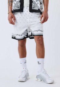 Pantalones cortos blancos con detalles negros, que presentan un patrón decorativo y un texto a lo largo del dobladillo. Combinados con calcetines blancos y zapatillas de deporte gris claro.